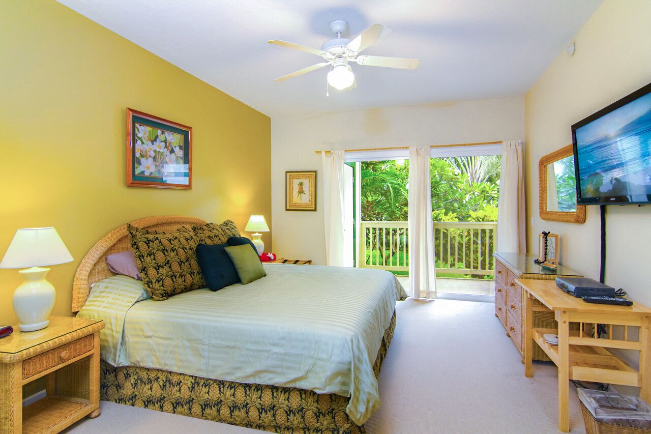 Une chambre à coucher comprenant un lit, des lampes de chevet, un ventilateur de plafond, une télévision, un bureau et un balcon avec vue sur la verdure.