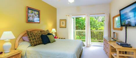 Une chambre à coucher comprenant un lit, des lampes de chevet, un ventilateur de plafond, une télévision, un bureau et un balcon avec vue sur la verdure.