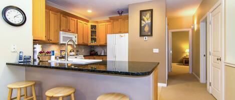 A kitchen with wooden cabinets, a black countertop, a refrigerator, and a clock on the wall.