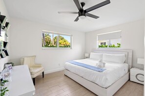Une chambre à coucher moderne avec un grand lit, un ventilateur de plafond, une table de chevet, une chaise, une fenêtre donnant sur des palmiers et un miroir fixé au mur.