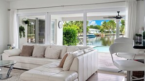 A modern living room with a white sofa, a glass coffee table, and a view of a pool and tropical landscape.