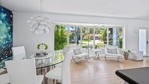 A modern living room with a glass dining table, white chairs, a white sofa, and a large window with a view of greenery and buildings.