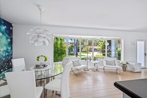 A modern living room with a glass dining table, white chairs, a white sofa, and a large window with a view of greenery and buildings. - Waterfront Home: Near Beach, Pool, Dock, Best View (Fort Lauderdale)