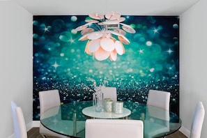 Un coin repas avec une table ronde en verre, des chaises blanches et une fresque murale représentant un ciel étoilé.