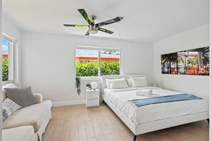 Een moderne slaapkamer met een wit bed, een plafondventilator, een raam met uitzicht op groen en een schilderij aan de muur.