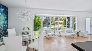 A modern living room with a glass dining table, white chairs, a white sofa, and a large window with a view of greenery and buildings.