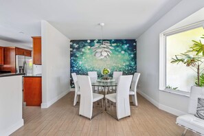 Een moderne eethoek met een glazen tafel en witte stoelen, een decoratief wandtapijt en een groot raam met een plant.