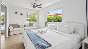 A bedroom with a large bed, a ceiling fan, two windows with views of greenery, a white sofa, and a small table with a plant.