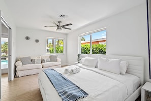 A bedroom with a large bed, a ceiling fan, two windows with views of greenery, a white sofa, and a small table with a plant. - Waterfront Home: Near Beach, Pool, Dock, Best View (Fort Lauderdale)