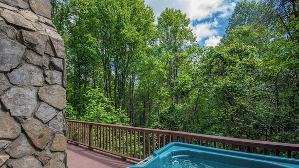 Un bain à remous sur une terrasse en bois, entouré d’un mur de pierres, avec une zone boisée en arrière-plan.