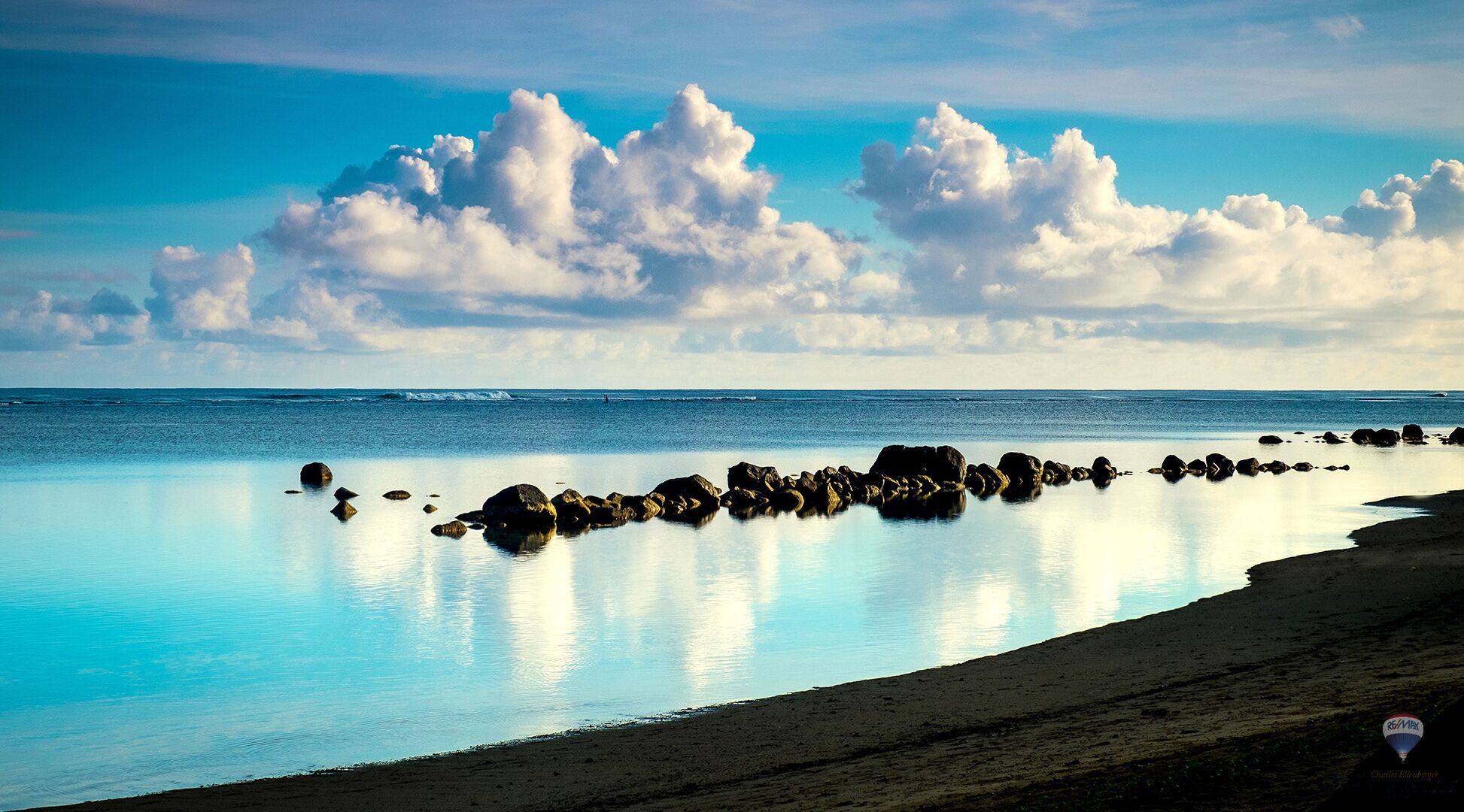 Una playa con agua tranquila, rocas dispersas y un cielo despejado con nubes.