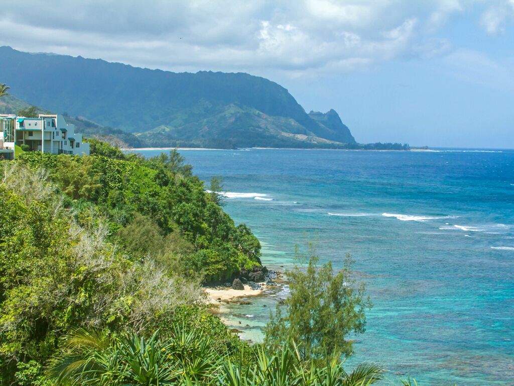 A coastal view with a building on a hillside, clear blue sea, and lush greenery.