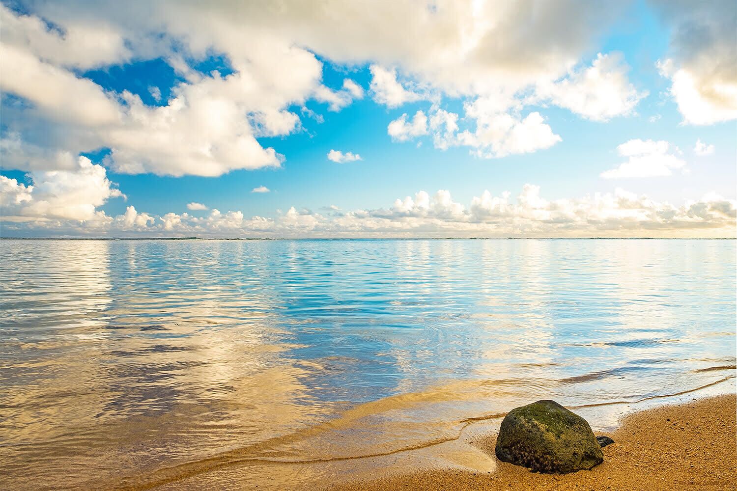 A beach with clear water reflecting the sky, a large rock in the foreground, and a distant shoreline.