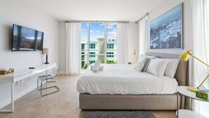 A modern bedroom with a large bed, a desk, and a view of buildings through the window. - Hemingway Penthouse South - Two Blocks from the Beach! (Deerfield Beach)