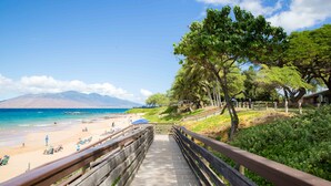 A wooden boardwalk leading to a beach with clear blue water and distant mountains.
