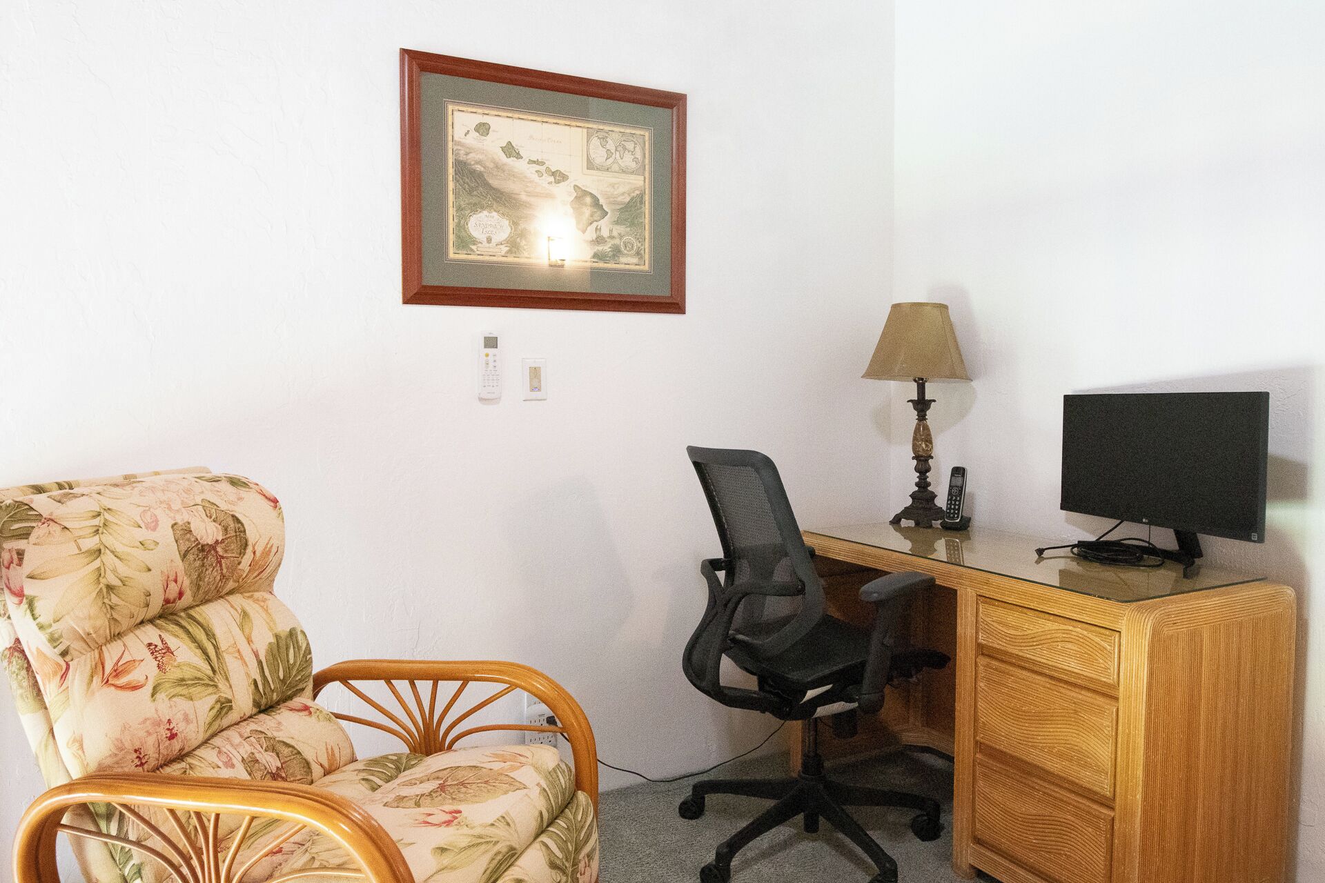 A room with a wooden desk, a chair, a lamp, a framed map, and a bed with floral upholstery.