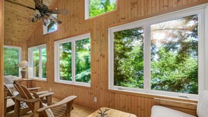 Interior de una cabaña de madera con ventanas amplias, ventilador de techo y muebles rústicos.