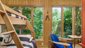 Una cabaña de madera con literas, una mecedora y una mesa junto a un ventanal grande con vistas a árboles.