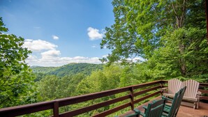 A wooden deck with a railing, green Adirondack chairs, and a view of a forested mountain range.