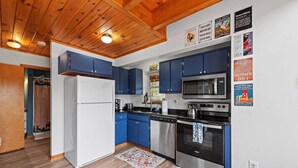 A kitchen with blue cabinets, a white refrigerator, a microwave, and an oven. There are posters on the wall and a wooden ceiling.