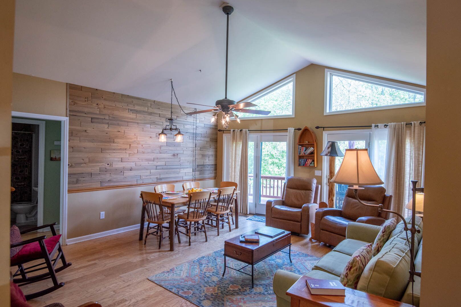 A spacious living room with a wooden wall, a dining area with wooden chairs, a sofa, a coffee table, and a ceiling fan.