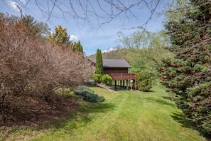 A wooden cabin with a deck, surrounded by a well-maintained lawn and various trees. - River Haven on the New River | Ultimate Outdoor Living | Hot Tub, Ping Pong, & More (Fleetwood)