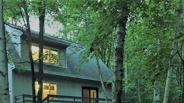 A two-story cabin with a wooden deck and a stack of firewood in front.