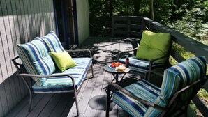 A wooden deck with a sofa and two chairs, a small table with a drink and snacks, and a view of a forested area.