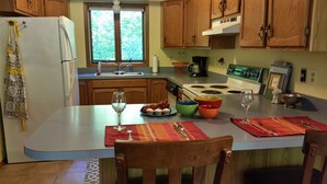 A kitchen with wooden cabinets, a white refrigerator, a window with a view of trees, and a dining area with a table set for two.