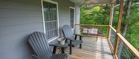 A wooden deck with two Adirondack chairs and a small table, overlooking a lush green forest.