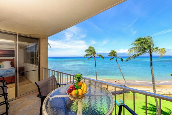 Un balcón con una mesa de vidrio y un frutero, con vista a una playa con palmeras y agua azul cristalina.