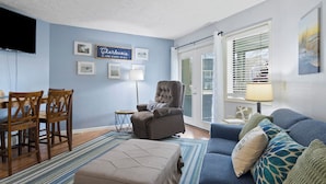 A living room with a blue wall, a television, a chair, a sofa, and a window with blinds.