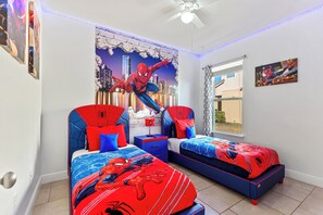 A bedroom with two beds, each with a Spider-Man themed bedspread, a cityscape mural on the wall, and a ceiling fan.