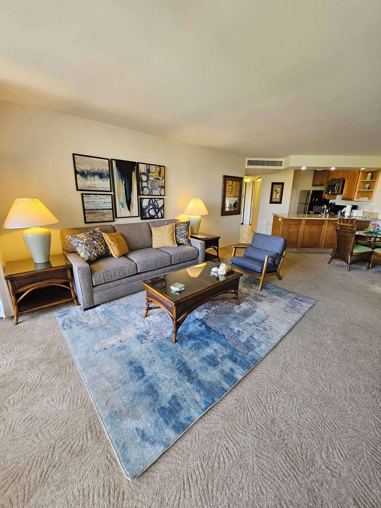 A living room with a sofa, two armchairs, a coffee table, and a rug. There are two lamps, a kitchen in the background, and artwork on the wall.