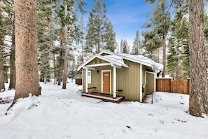 Une petite cabane en bois dotée d’un porche, entourée d’un paysage enneigé et de grands pins.