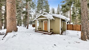 Una cabaña pequeña con puerta de madera y un porche, rodeada de árboles altos y cubierta de nieve.