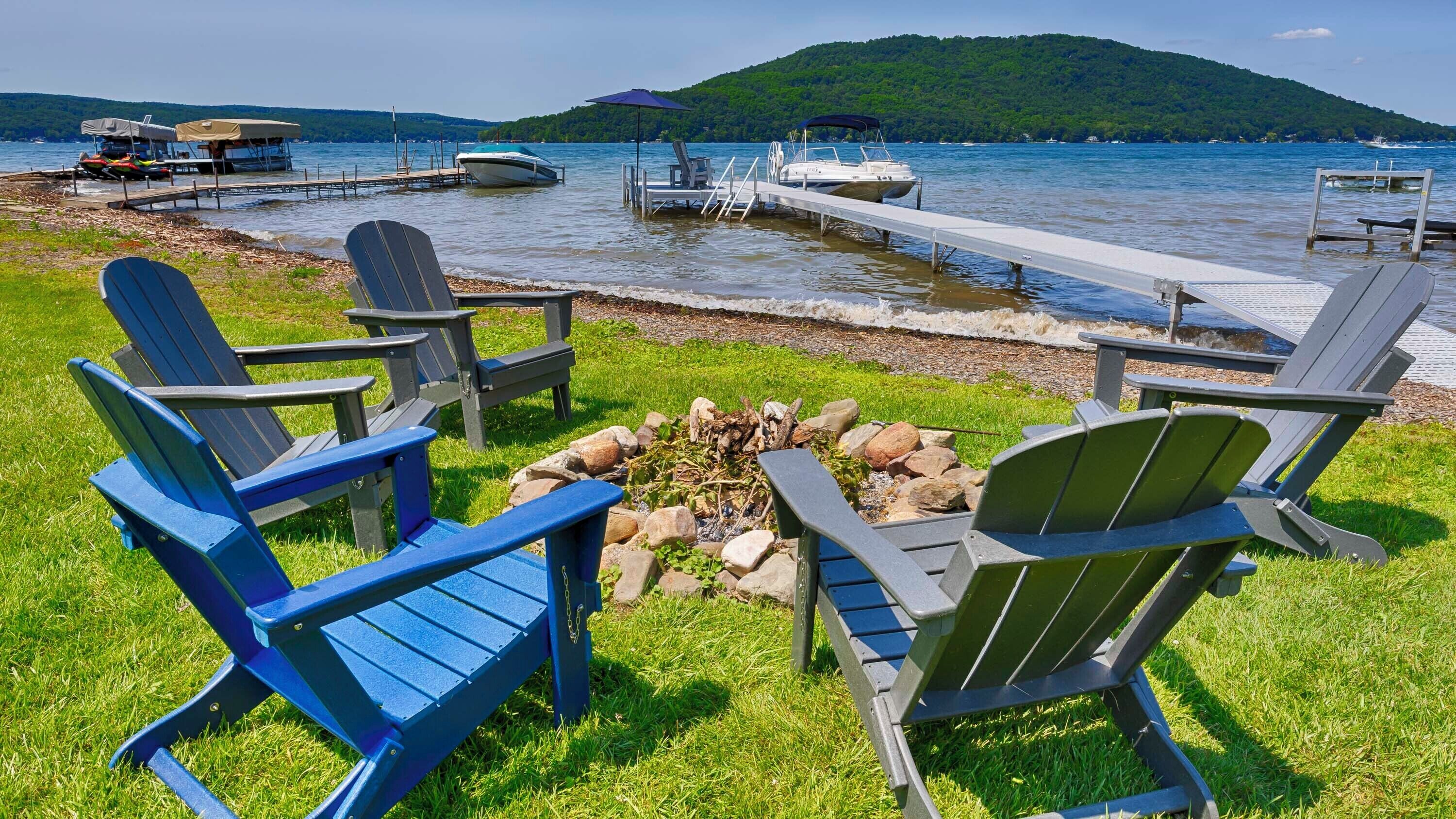 A lakeside setting with a fire pit surrounded by Adirondack chairs, a dock with boats, and a forested hill in the background.