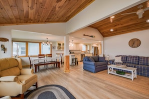 A spacious living area with a wooden ceiling, a dining table with chairs, a blue sofa, and a kitchen in the background.