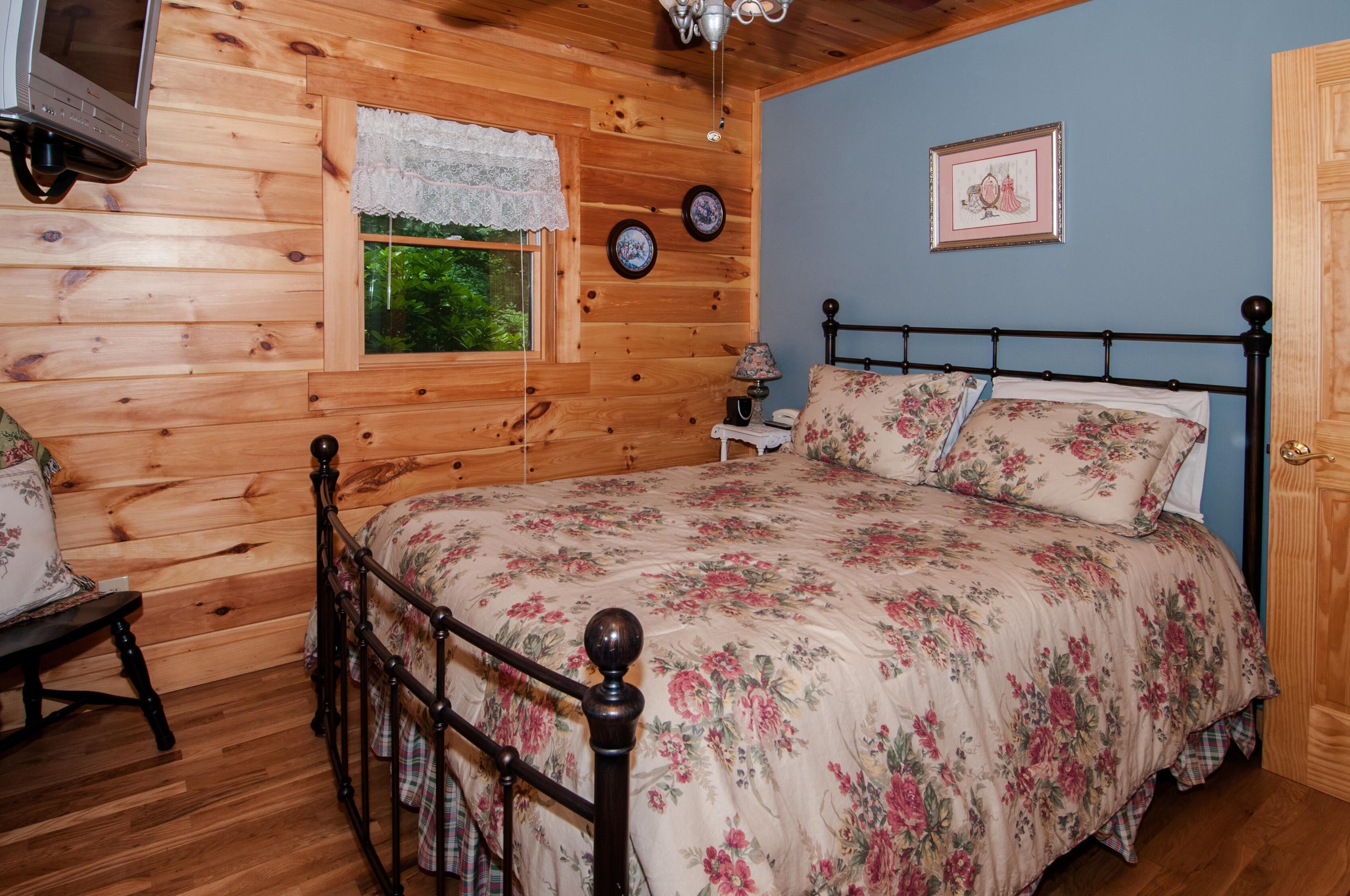 A wooden cabin bedroom with a bed, a window with lace curtains, a bedside table with a lamp, and a framed picture on the wall.