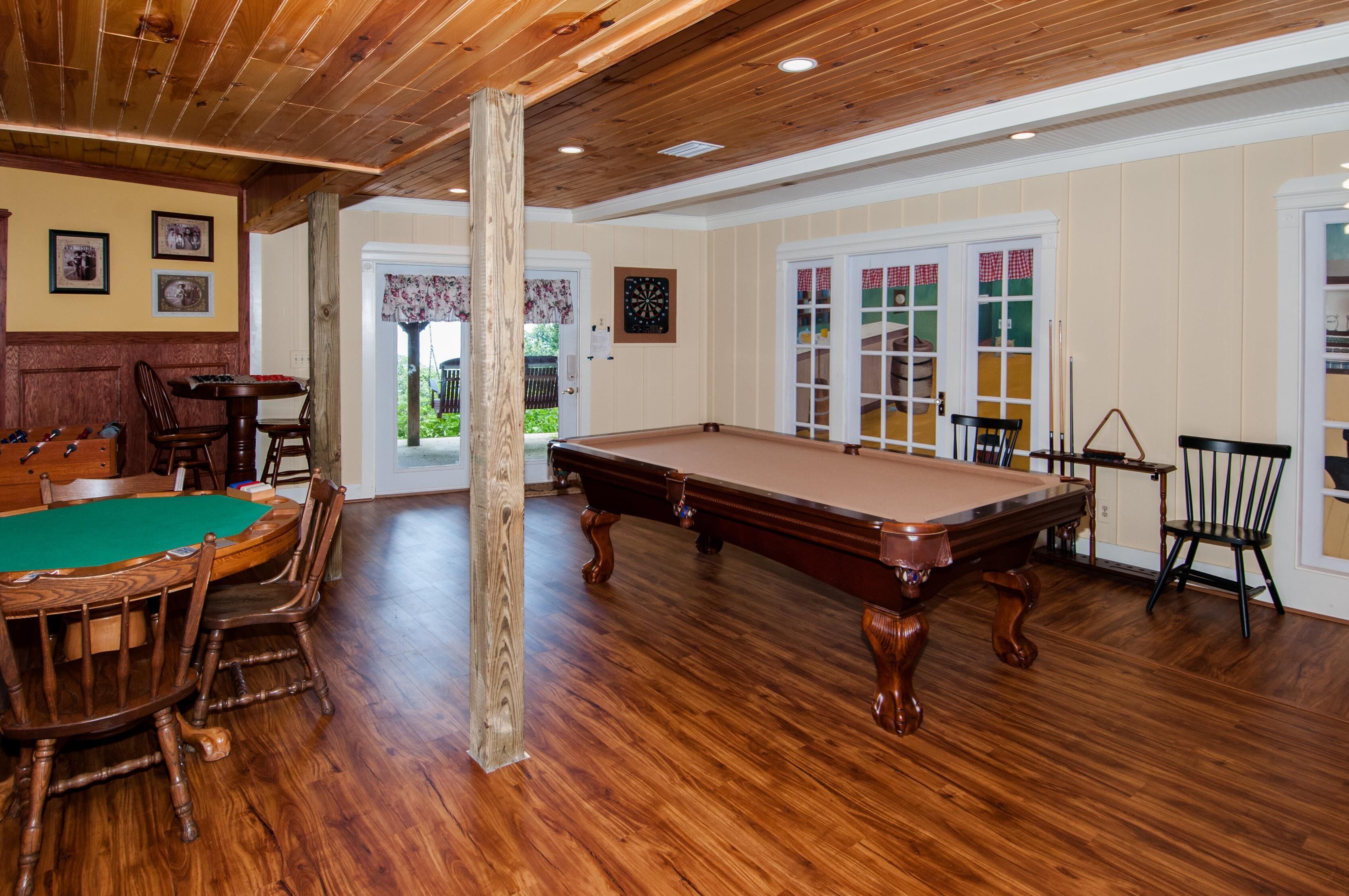 A pool table with surrounding wooden chairs and a dartboard in a room with wooden floors and a wooden ceiling.