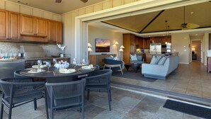 A spacious living area with a dining table set for six, surrounded by dark wicker chairs. A kitchen with wooden cabinets and a built-in stove is visible in the background. - Local Luxury Living! (Kamuela)