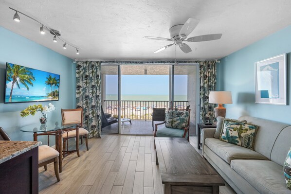 A living room with a balcony, a ceiling fan, and a large window with a view of the beach.