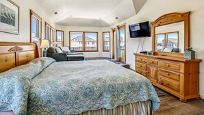 Een ruime slaapkamer met een groot bed, een houten dressoir, een televisie en grote ramen met uitzicht op huizen.