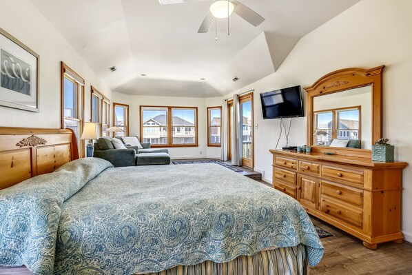 A spacious bedroom with a large bed, a wooden dresser, a television, and large windows offering a view of houses.