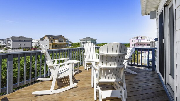 Een houten terras met witte Adirondack-stoelen en een uitzicht op huizen in de verte.