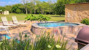 A hot tub surrounded by lounge chairs, a stone wall, and a grassy area with trees. - WW C302 Gracelanda (New Braunfels)