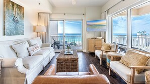 A spacious living room with a large window offering a view of the ocean and buildings. There is a ceiling fan, a sofa, a coffee table, and a wicker chair.