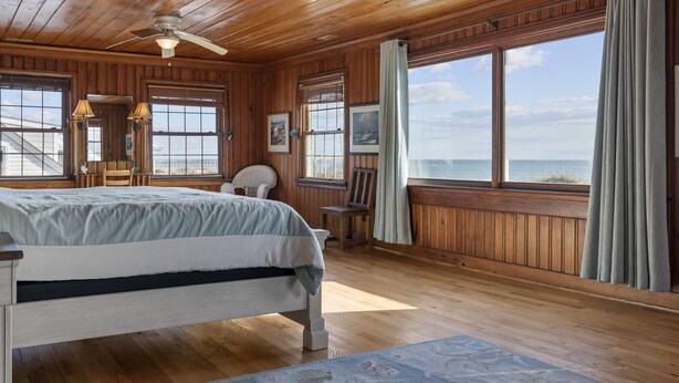 Une chambre située dans une cabane en bois, avec un lit, deux chaises, un bureau et une grande fenêtre donnant sur l’océan.