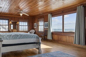 Een slaapkamer in een houten hutje met een bed, twee stoelen, een bureau en een groot raam met uitzicht op de oceaan.