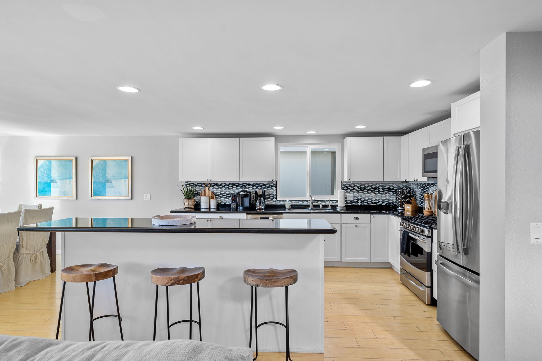 A modern kitchen with white cabinets, a black countertop, and stainless steel appliances. There are three wooden stools at the island, and a large window provides natural light.